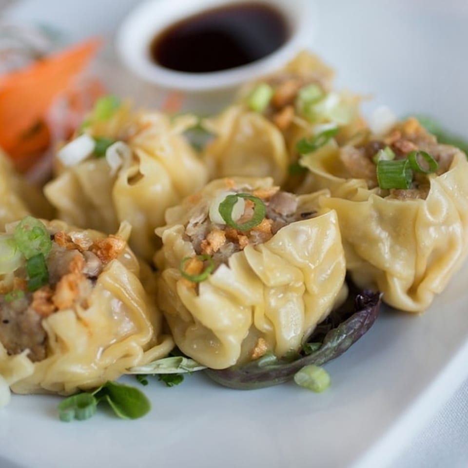 Steamed dumplings topped with green onions and crispy garlic, served with a side of soy sauce.