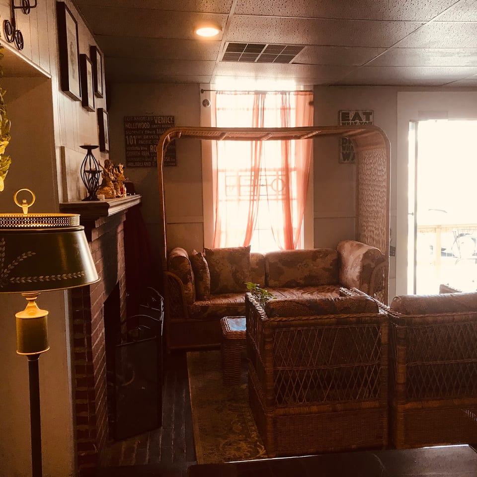 Cozy restaurant interior with wicker furniture, a fireplace, and warm lighting from a window.