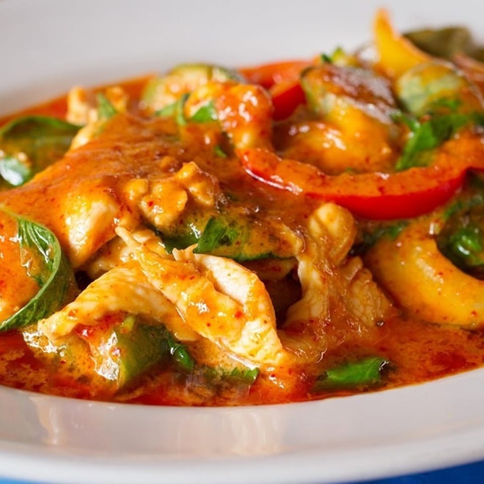 Close-up of a vibrant red curry dish with chicken, bell peppers, and basil in a white bowl.