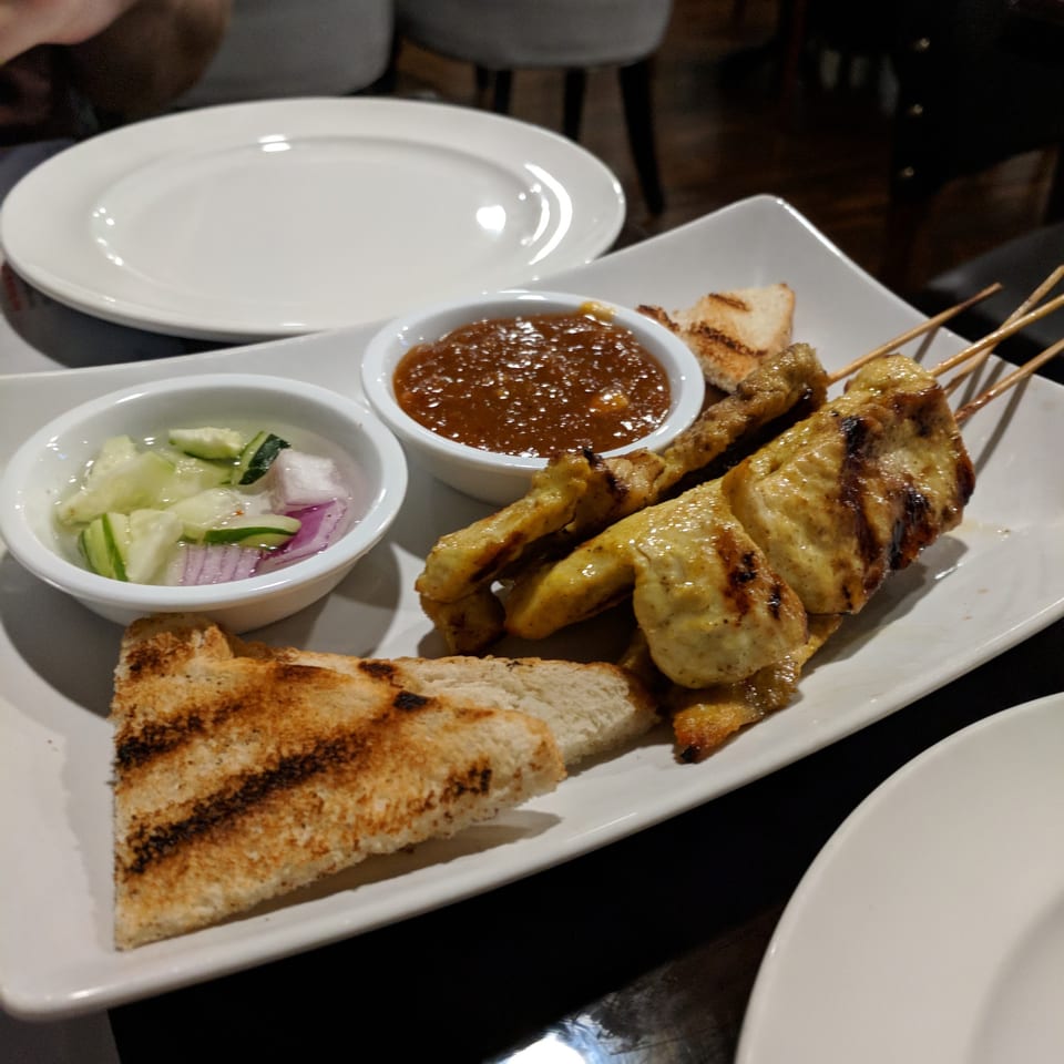 Chicken satay skewers with peanut sauce, cucumber salad, and grilled bread on a white plate.