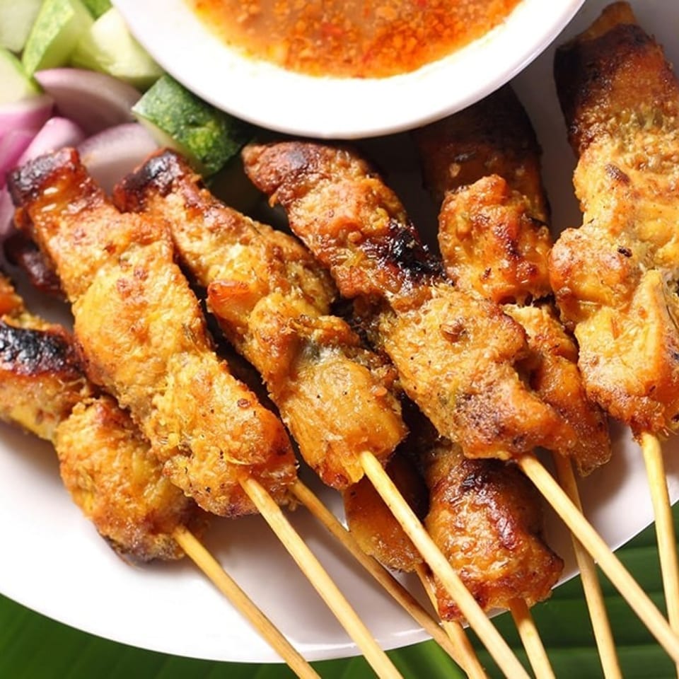 Grilled chicken satay skewers with peanut sauce, sliced cucumbers, and red onions on a white plate.